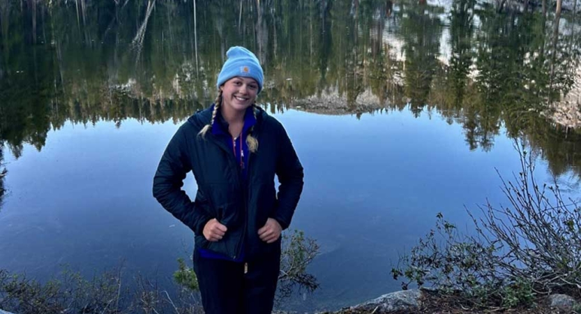 A person stands and smiles in front of a body of water that is reflecting the trees and hills that surround it. 